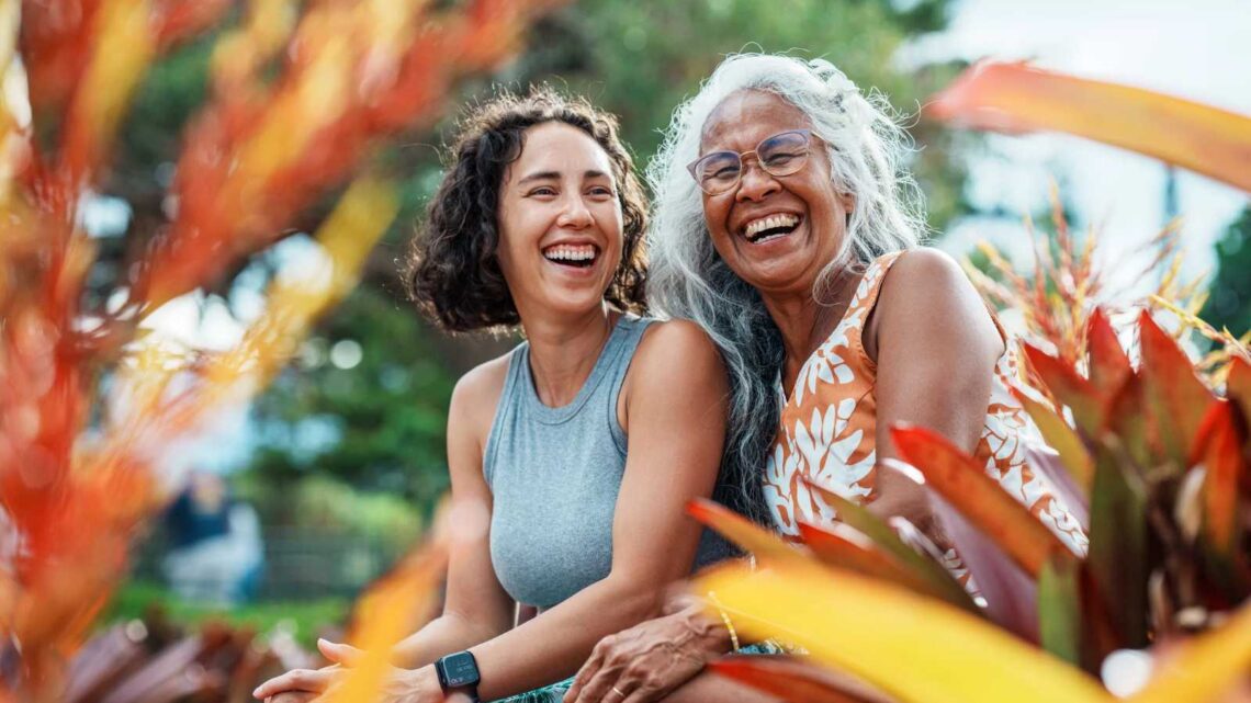 Two women sit outdoors, laughing and surrounded by plants with red and yellow leaves. The woman on the left has short curly hair and wears a gray sleeveless top. The woman on the right has long gray hair, glasses, and a patterned orange dress.