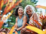 Two women sit outdoors, laughing and surrounded by plants with red and yellow leaves. The woman on the left has short curly hair and wears a gray sleeveless top. The woman on the right has long gray hair, glasses, and a patterned orange dress.